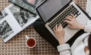 Kobieta pisze na laptopie, obok leży otwarta księga i filiżanka herbaty. Zastanawia się, ile kosztuje artykuł sponsorowany.