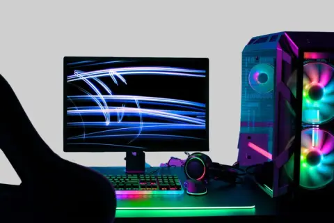 Gracz wpatrzony w monitor z niebieskimi smugami światła. Obok komputer z kolorowymi wentylatorami. Idealne miejsce na fajne słowa po angielsku na nick.