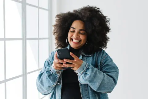 Uśmiechnięta kobieta z burzą loków przegląda telefon, szukając inspiracji, jak udostępnić czyjąś relację na Instagramie.