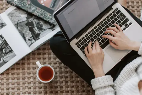 Kobieta pisze na laptopie, obok leży otwarta księga i filiżanka herbaty. Zastanawia się, ile kosztuje artykuł sponsorowany.