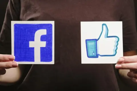 Dwie ręce trzymają kartki z logo Facebooka i kciukiem w górę.