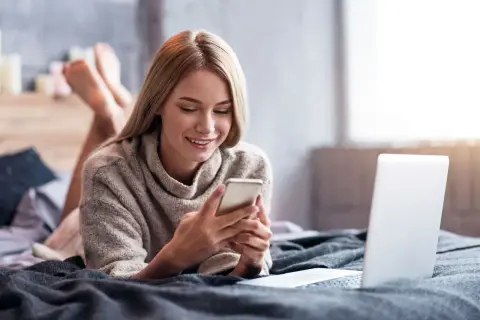 Uśmiechnięta dziewczyna leży na łóżku z telefonem w dłoniach, szukając informacji, jak doładować komuś konto w Play z karty na kartę. Obok leży laptop.