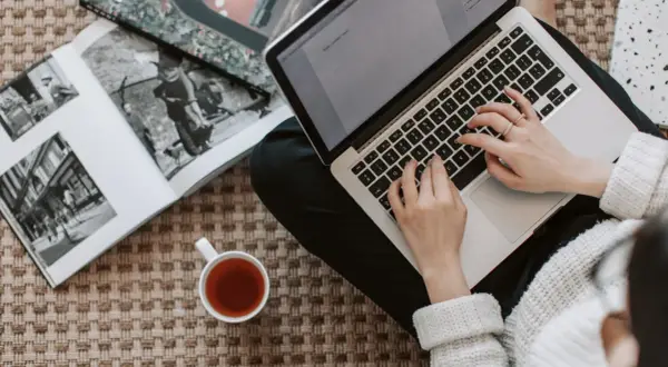 Kobieta pisze na laptopie, obok leży otwarta księga i filiżanka herbaty. Zastanawia się, ile kosztuje artykuł sponsorowany.