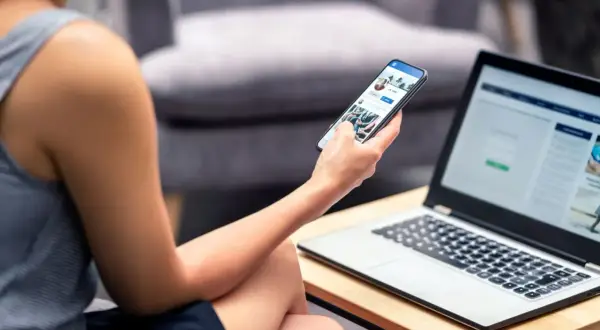 Kobieta przegląda telefon, szukając informacji, jak odzyskać usunięte konto na FB. Obok leży laptop.