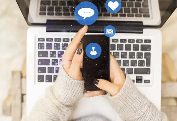 Dłonie trzymające smartfon nad klawiaturą laptopa. Ikony czatu, serca i maila unoszą się w powietrzu. Czy jak usunę czat na Messengerze to druga osoba to widzi?