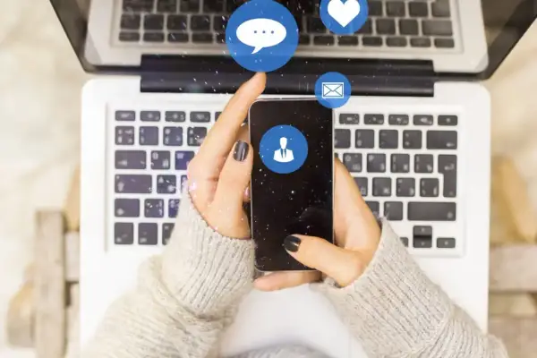 Dłonie trzymające smartfon nad klawiaturą laptopa. Ikony komunikatorów unoszą się w powietrzu. Zastanawiasz się, dlaczego na Messengerze nie widać, kiedy ktoś był aktywny?